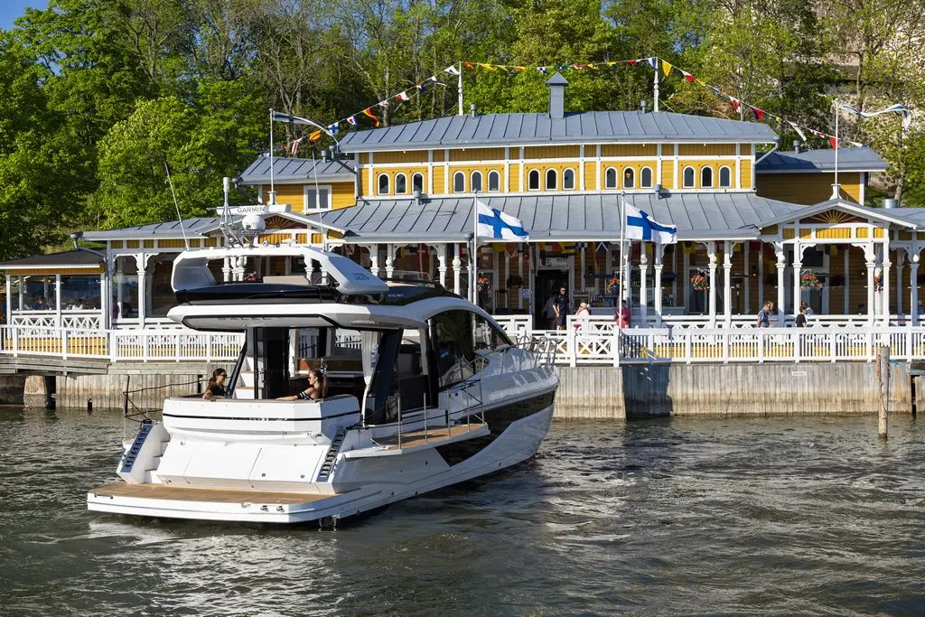 A luxury motorboat approaches a vibrant yellow waterfront restaurant with Finnish flags, set against lush greenery.
