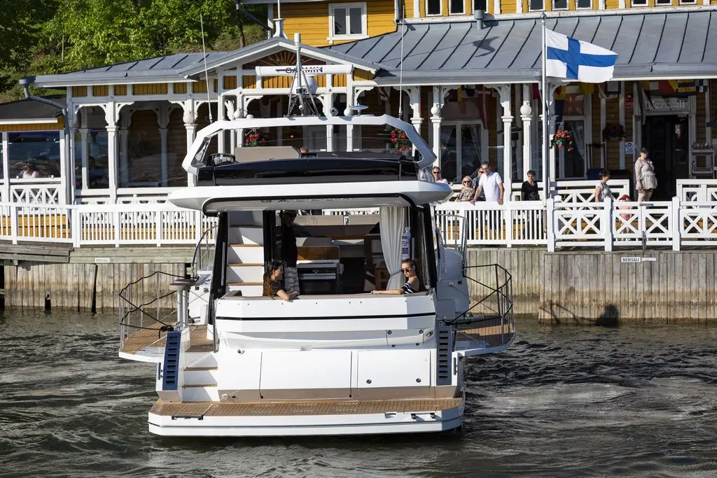 A motorboat approaches a waterfront restaurant, featuring a Finnish flag, with diners visible on the outdoor deck.