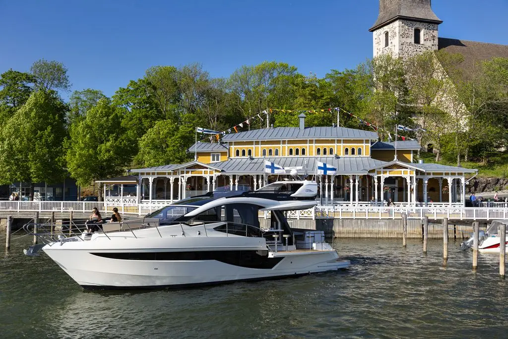Modern white yacht docked by a waterfront restaurant, flanked by lush trees and a historic church in bright sunlight.