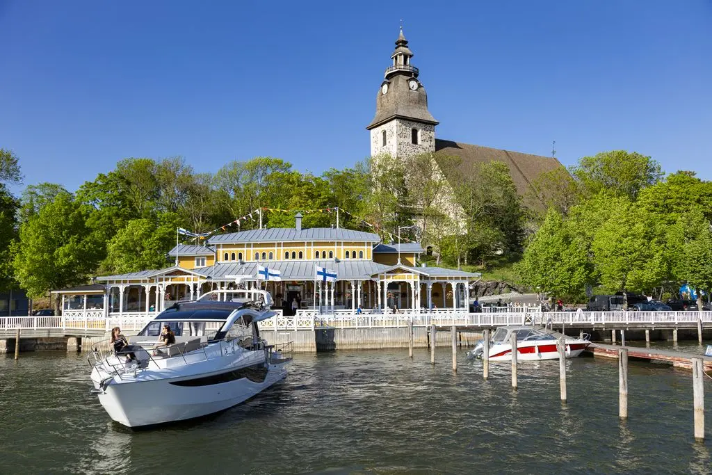 A scenic lakeside view featuring a boat, a waterfront restaurant with flags, and a church tower in the background.