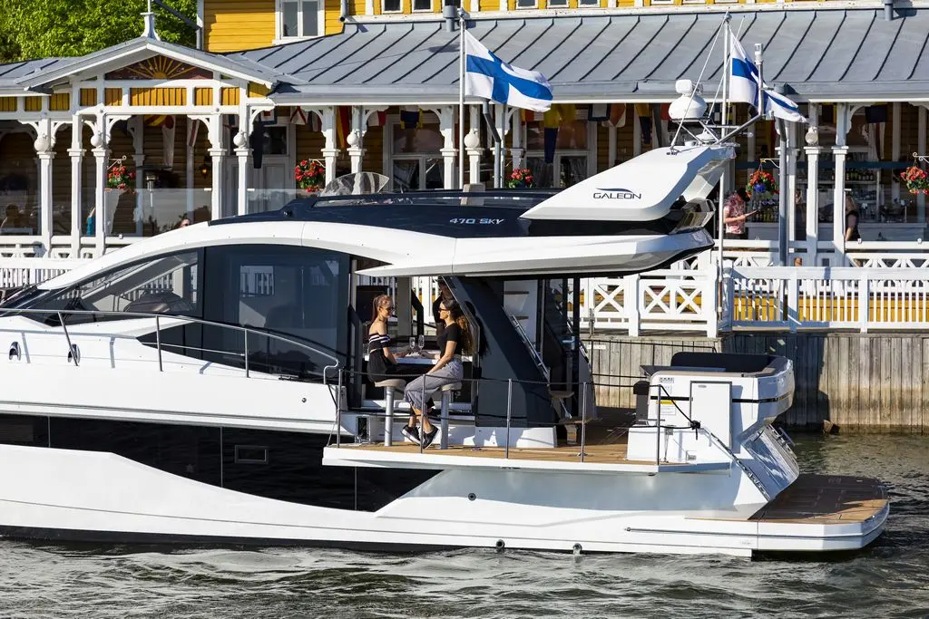 A sleek white yacht with two women onboard, docked near a vibrant waterfront building with flowers and flags.