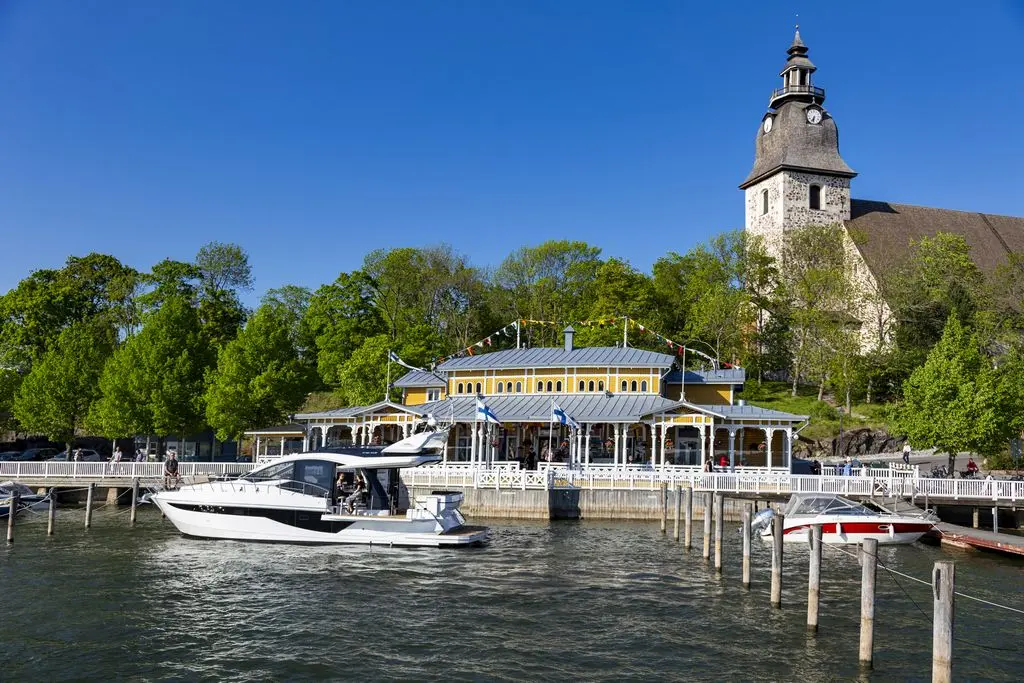 A white motorboat approaches a waterfront restaurant with a clock tower, surrounded by green trees and blue skies.