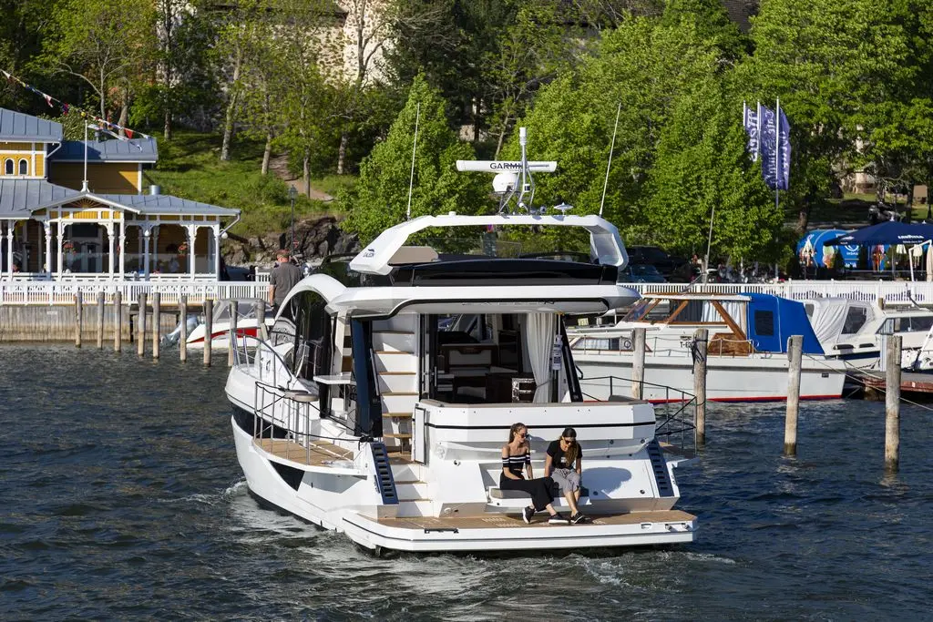 Two women sit on the deck of a luxury boat, moored near a waterfront restaurant and surrounded by trees and smaller boats.