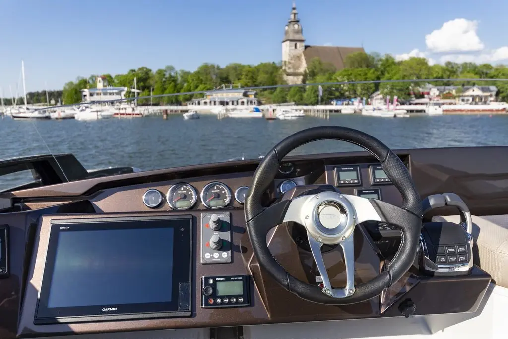 Boat steering wheel and dashboard with a GPS screen, overlooking a marina with boats and trees in the background.