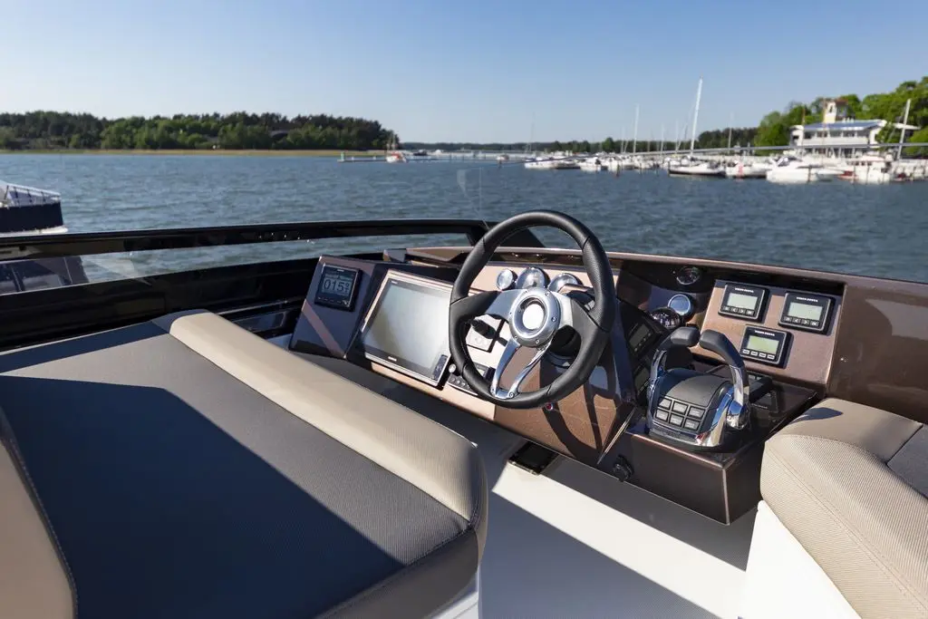 Cockpit of a luxury boat featuring a steering wheel, digital displays, and control panel, with a scenic water view in the background.