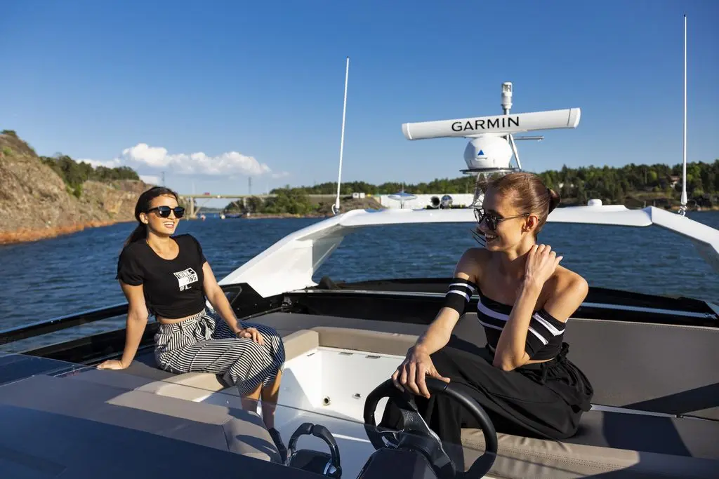 Two women on a boat enjoy the sunshine, one sitting elegantly and the other steering, with a Garmin device in view.
