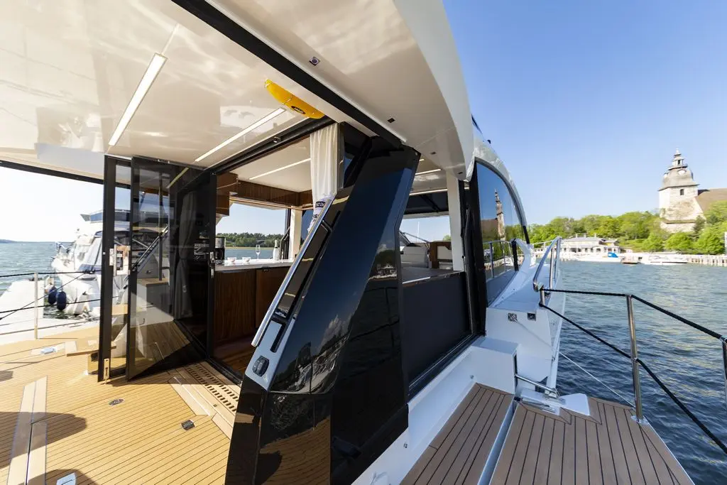Luxury yacht interior with open glass doors, wooden deck, scenic waterfront, and a historic building in the background.