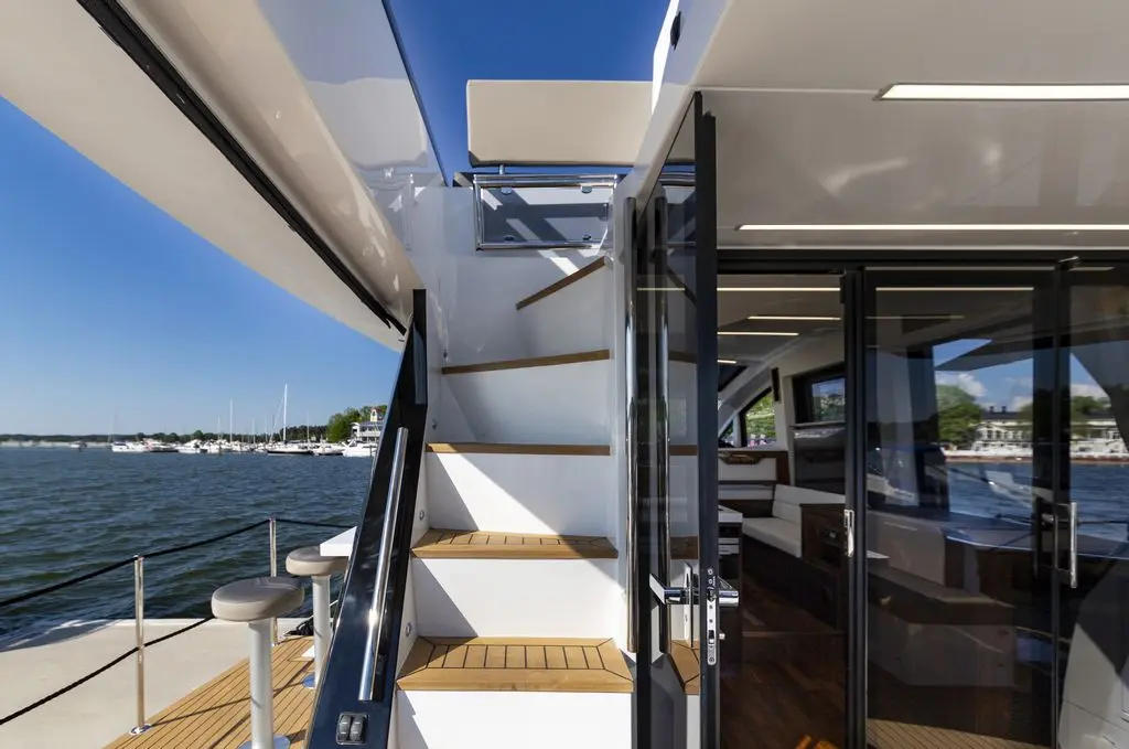 Modern yacht interior with glass doors, wooden stairs, and a view of a marina in the background under a clear blue sky.