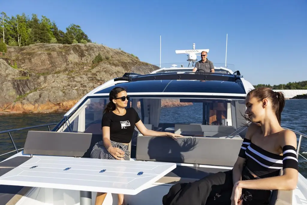 Two women relax on a boat, one wearing sunglasses and a black shirt, with a man steering in the background and scenic rocks visible.
