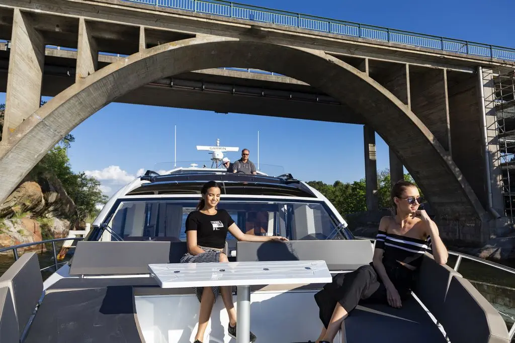 Three people relax on a boat under a bridge, with greenery and rocks visible in the background. A GARMIN device is on the boat.