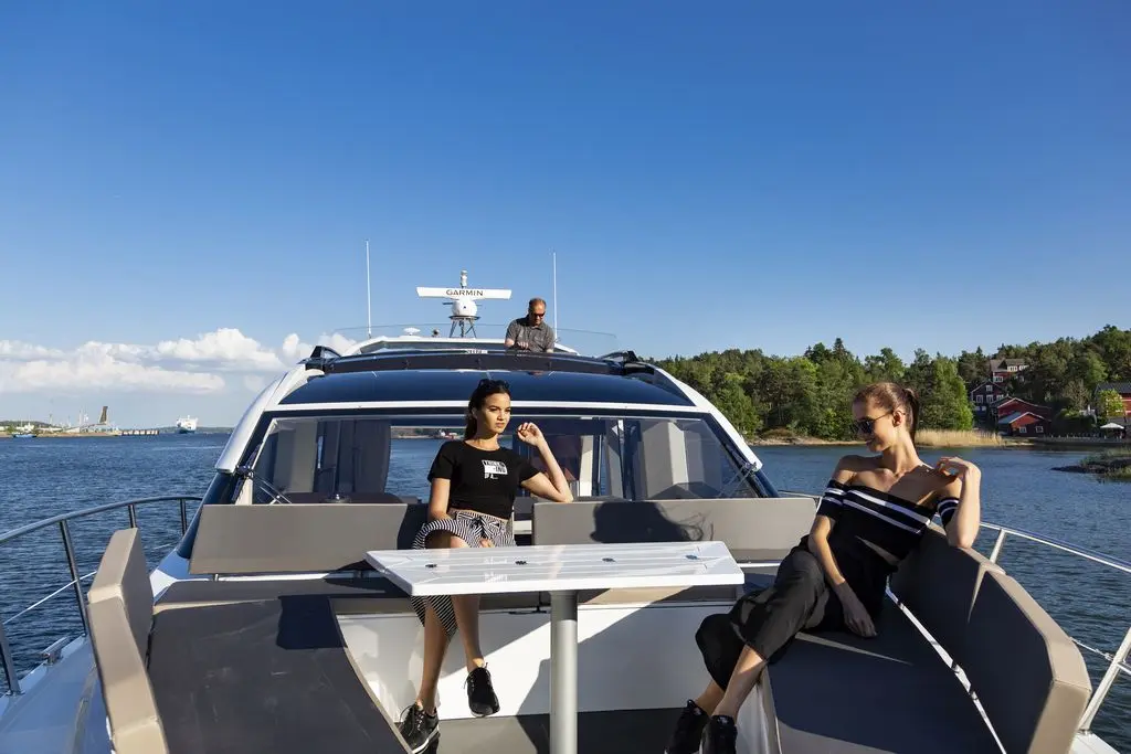 Two women relax on a yacht, with one sitting at a table and the other reclining, while a man steers the boat.