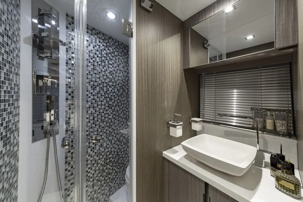 Modern bathroom with a glass shower, mosaic tiles, sink, and wooden cabinetry, featuring stylish toiletries and lighting.