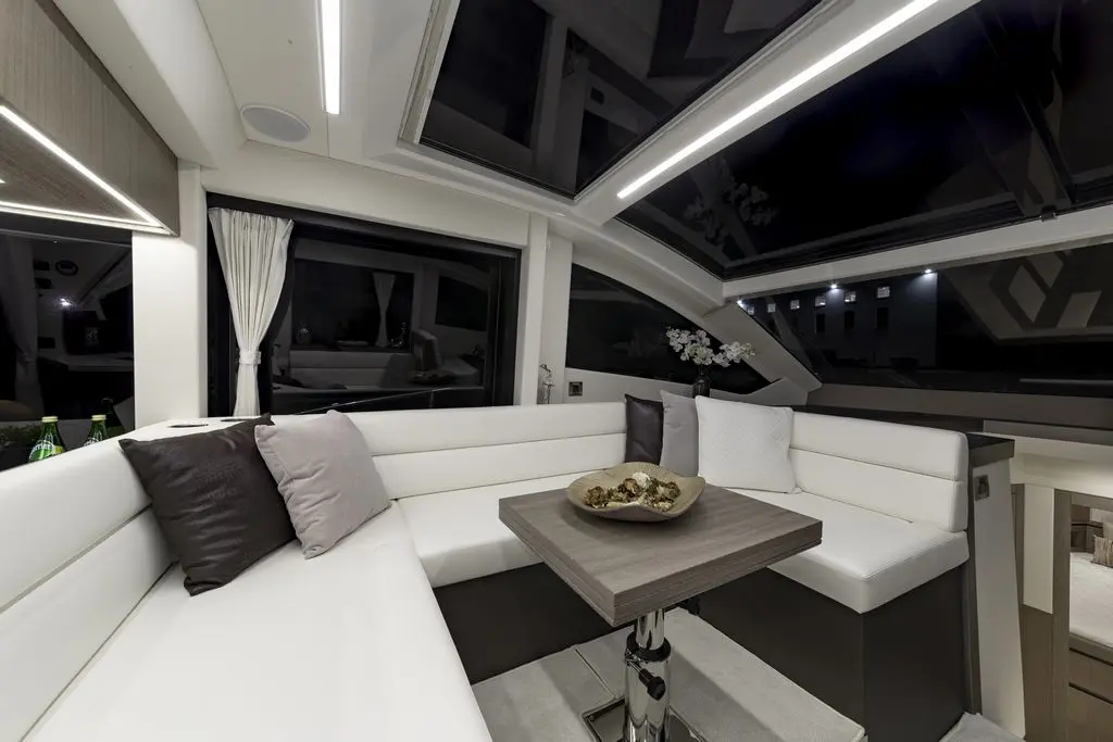 Interior of a modern yacht lounge featuring white seating, a wooden table, decorative cushions, and large windows with curtains.