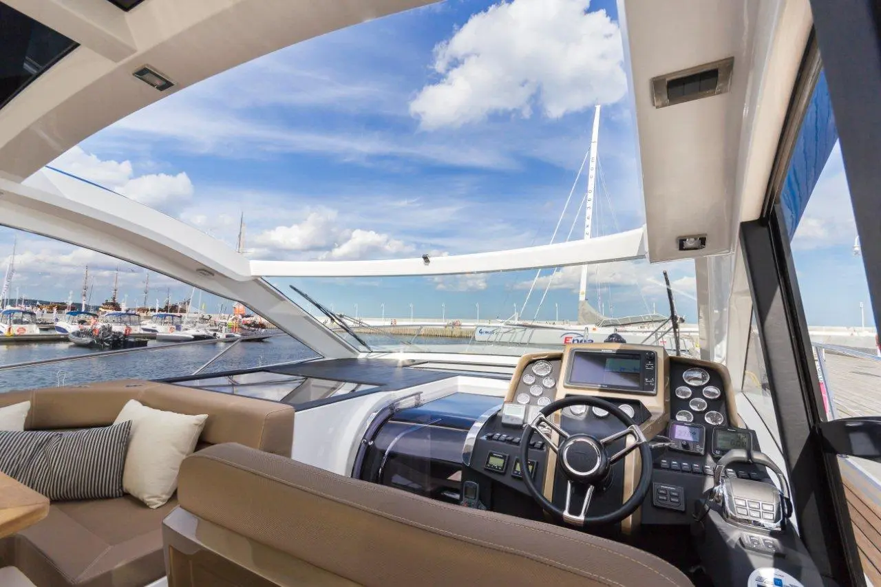Modern yacht interior with a navigational dashboard, plush seating, and a view of the marina under a blue sky.