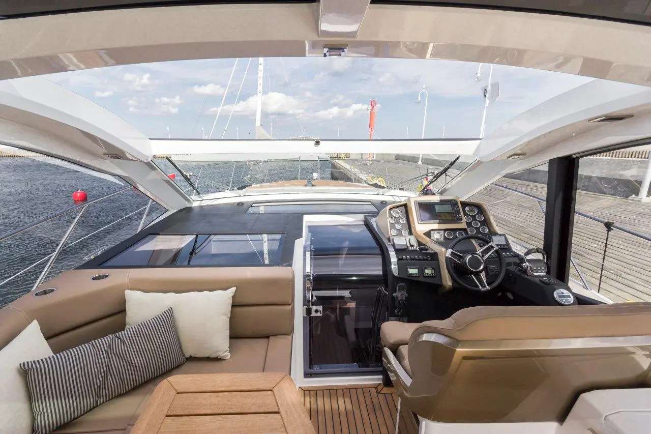 Luxury yacht interior with a cockpit view, dashboard, leather seating, and seating area, near a marina with boats.