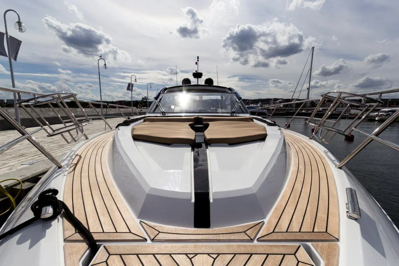 View of a luxury motorboat's front deck, featuring cushioned seating and wooden flooring, docked with a marina in the background.