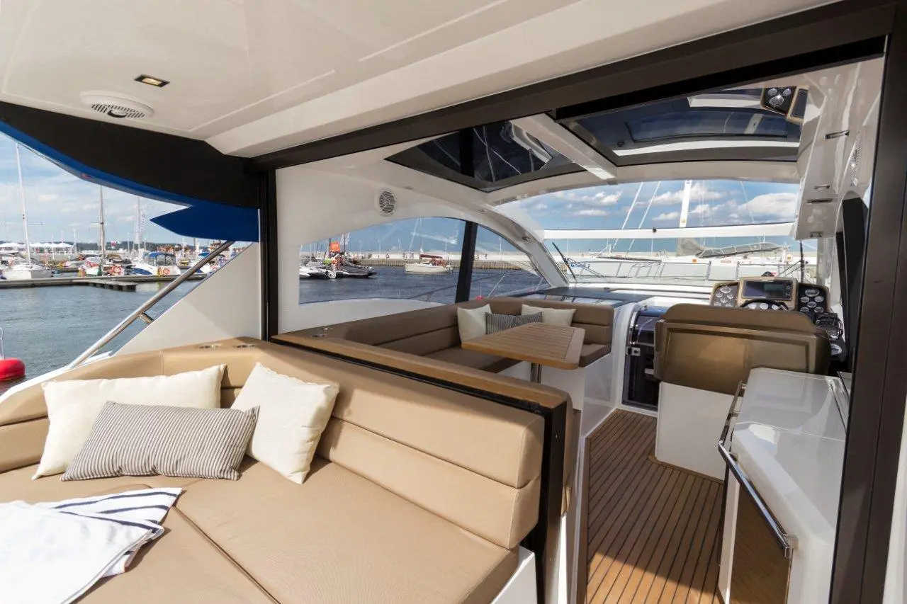 Interior of a modern yacht with beige seating, a wooden table, and a view of a marina filled with boats in the background.