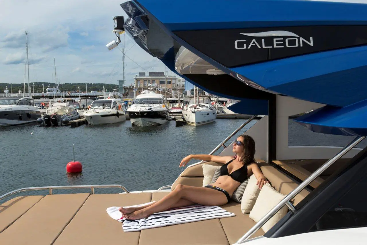 A woman in a black bikini relaxes on a yacht in a marina, surrounded by boats and the water, with a striped towel.