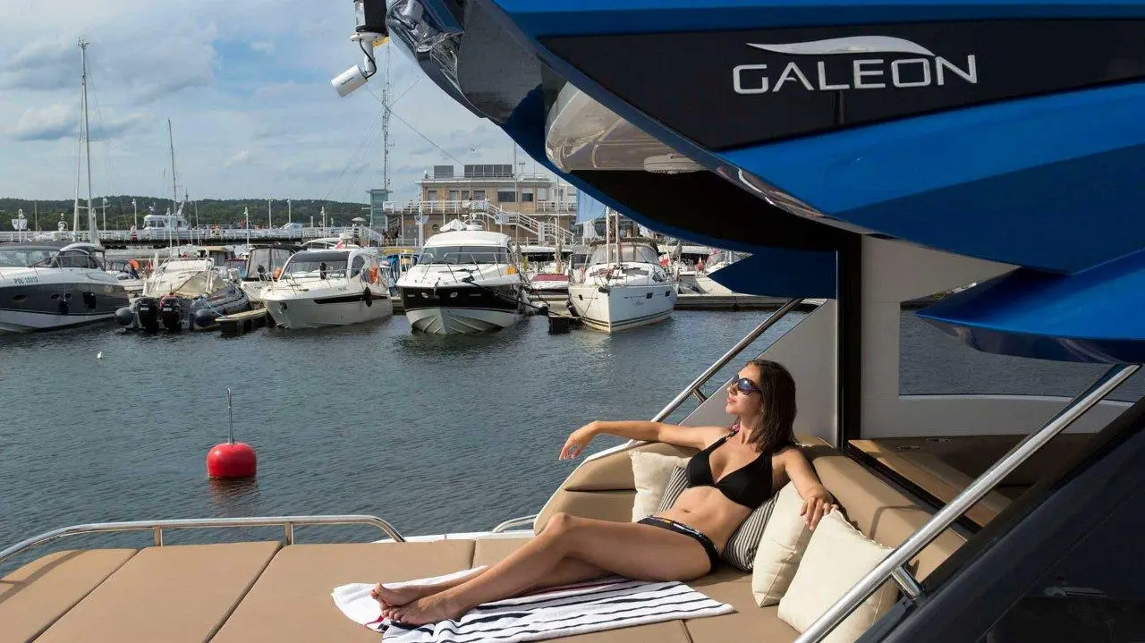 Woman in a bikini lounges on a yacht's deck, with boats and a marina in the background; 