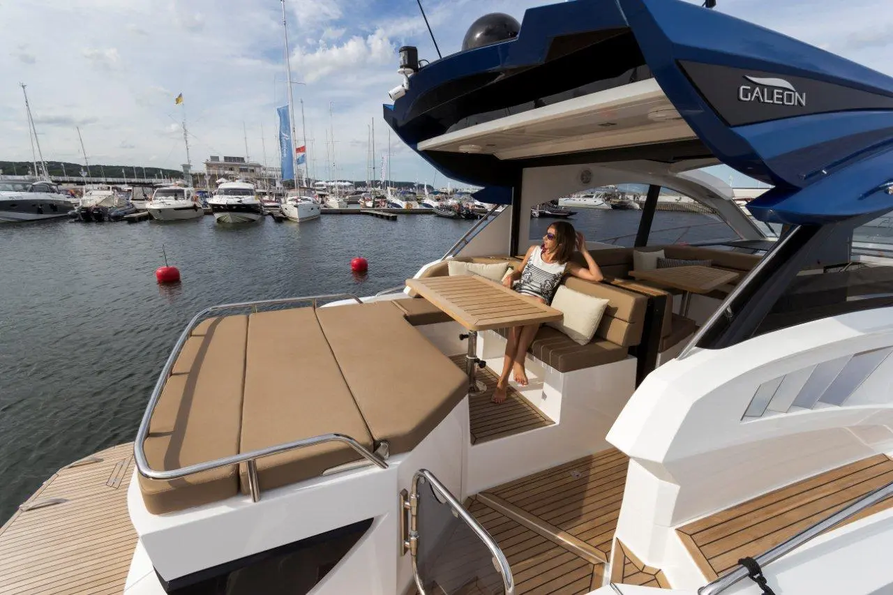 A woman relaxes on a yacht's deck with a table, overlooking a marina filled with boats and a blue sky.