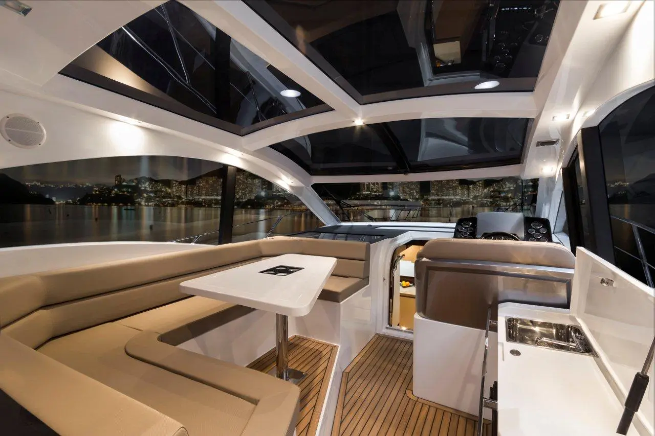 Interior of a modern boat featuring a cozy seating area, a table, and a helm with city lights visible through large windows.
