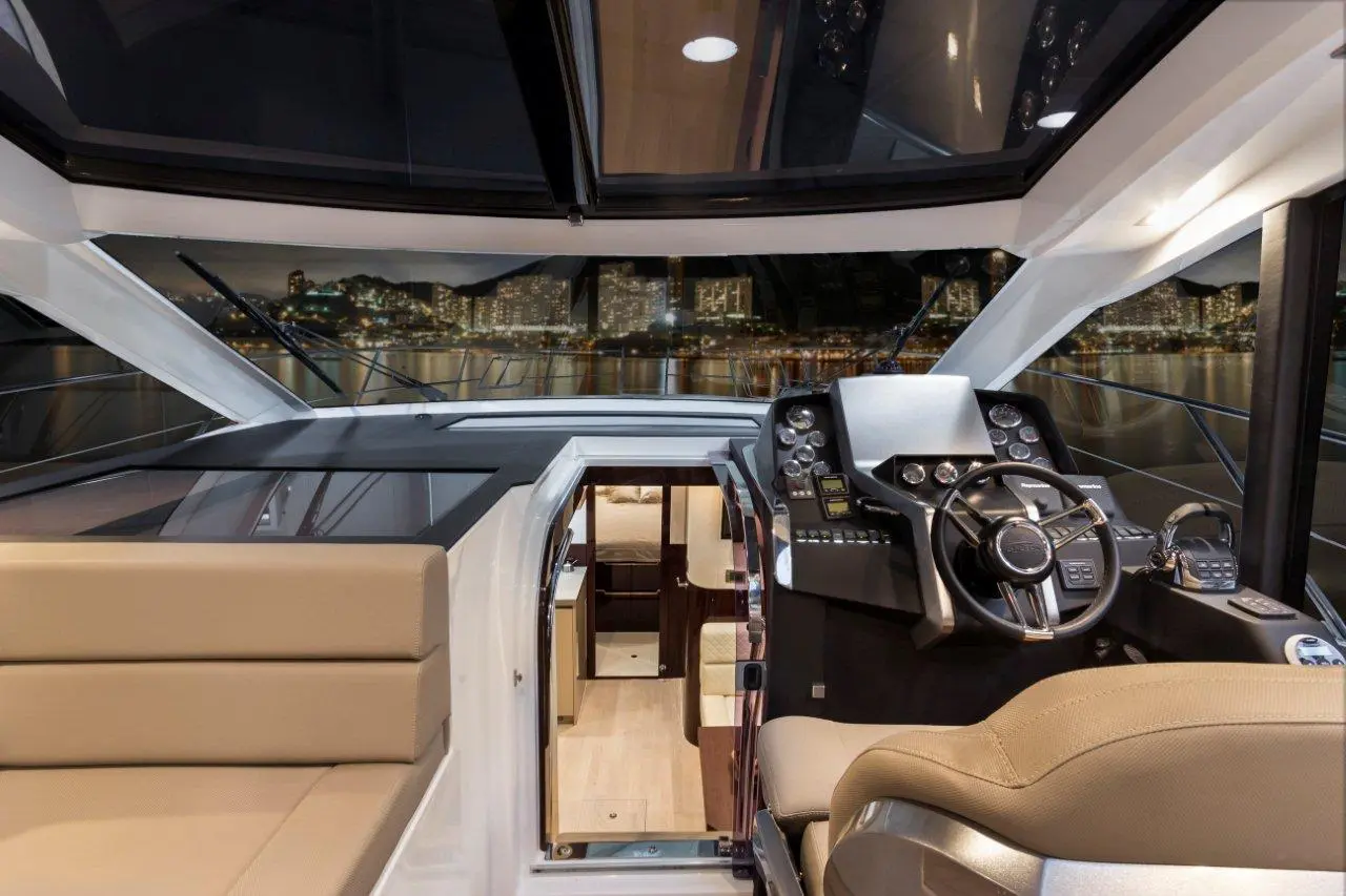 Modern yacht interior featuring a helm with navigation controls, plush seating, and a view of city lights through large windows.