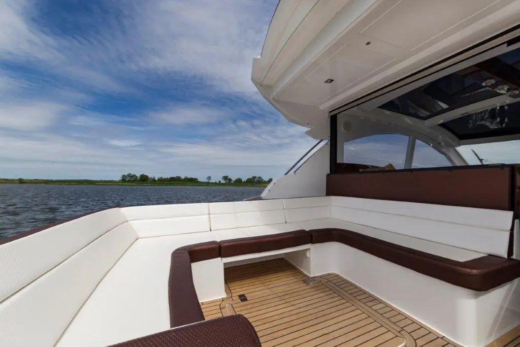 Luxurious boat interior featuring white and brown seating, wooden flooring, with a scenic view of a river and blue sky.