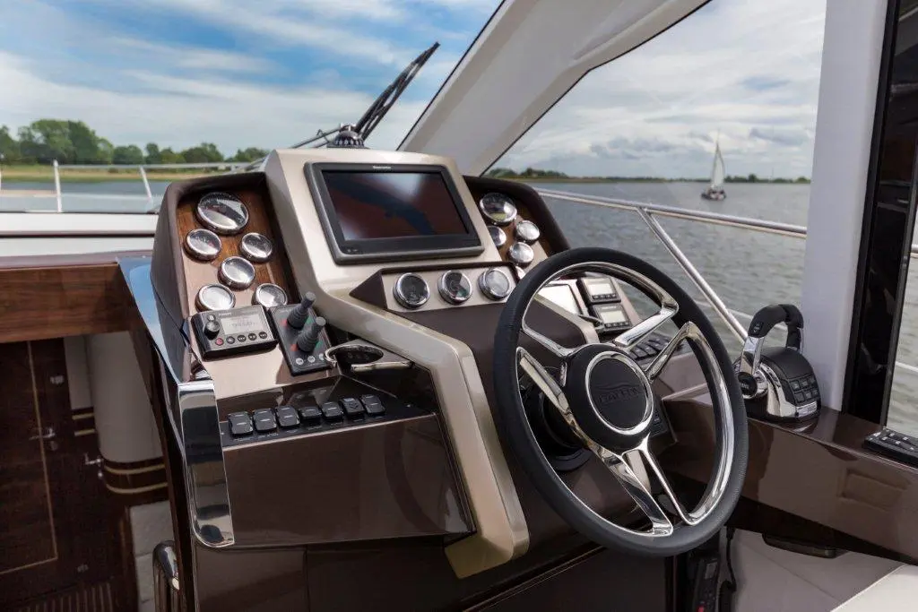 Boat cockpit with modern steering wheel, digital display, gauges, and controls, overlooking water and a sailboat in the background.