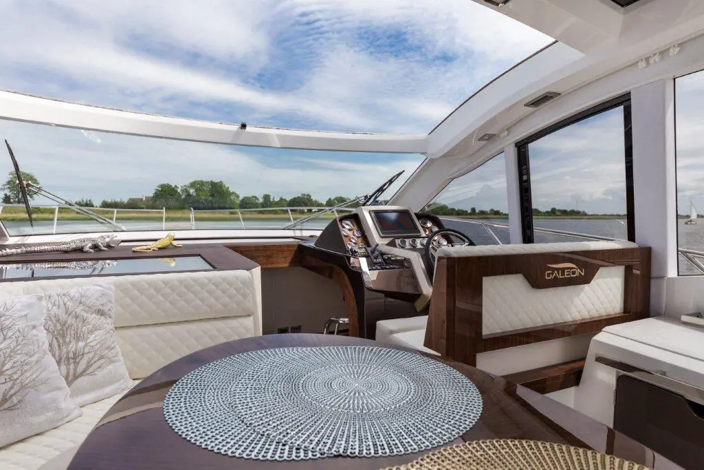 Luxury yacht interior featuring a steering console, plush seating, and a view of the waterway under a cloudy sky.