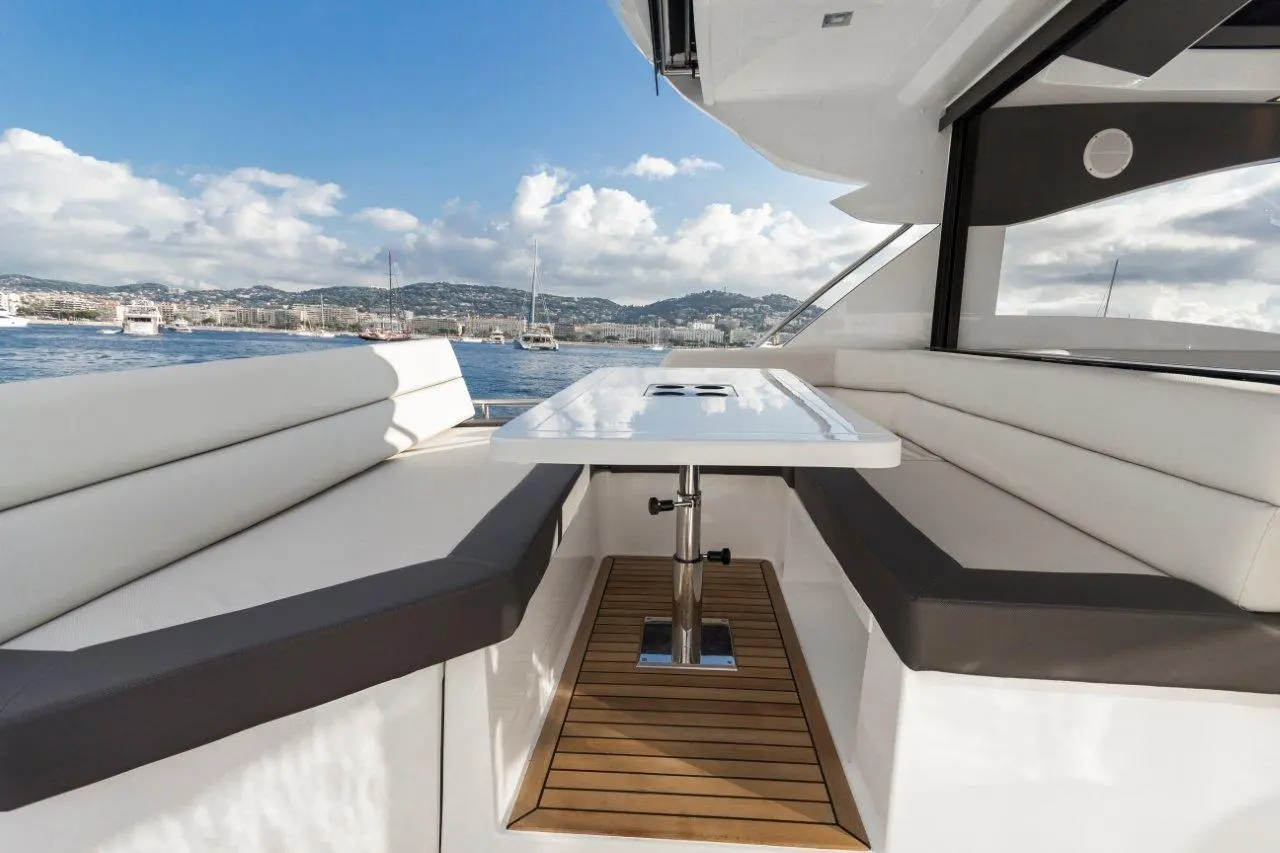 Modern yacht interior with white seating, a polished table, and scenic views of the coastline and boats.