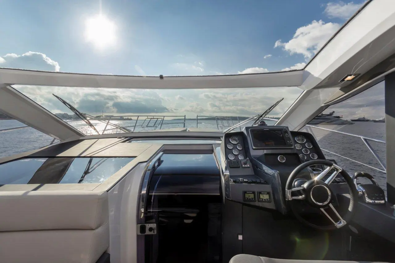 Interior view of a yacht's helm with a steering wheel and control panel, overlooking water under a bright sky.