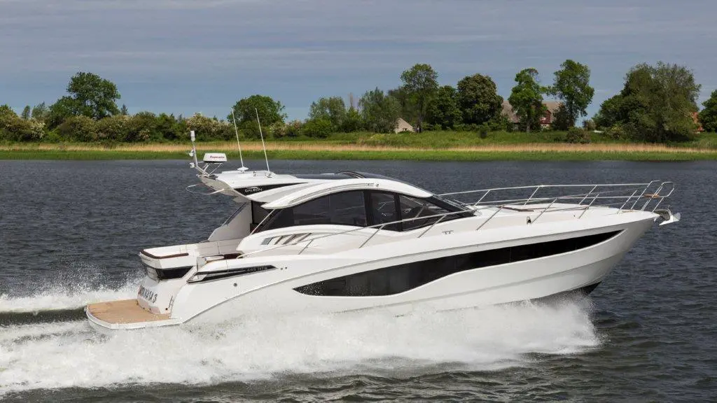 A sleek white motor yacht gliding through calm waters, leaving a foamy wake, with lush greenery in the background.