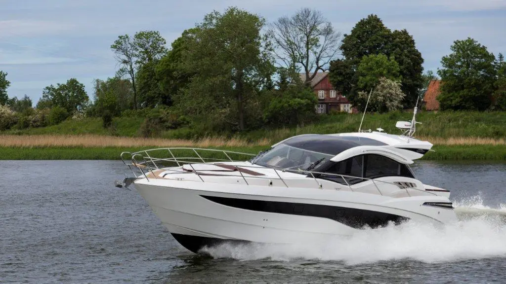 A sleek white motor yacht speeds through a calm river, with lush greenery and a house visible in the background.