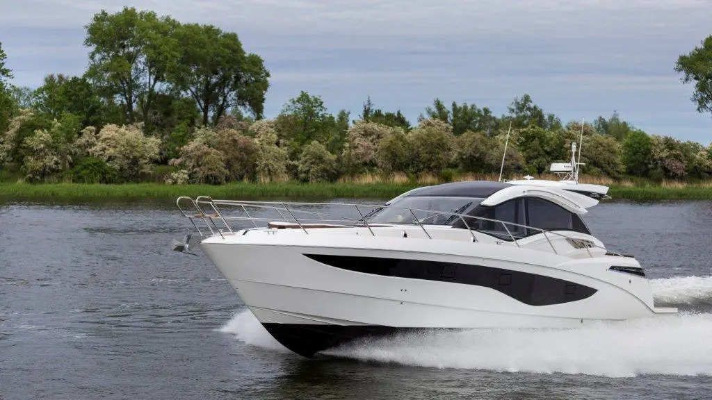 A sleek white motorboat speeds through calm waters, surrounded by lush greenery and trees under a cloudy sky.