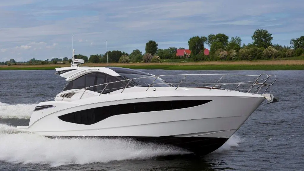 A sleek white yacht speeds across a calm river, surrounded by lush greenery and a distant red-roofed house.