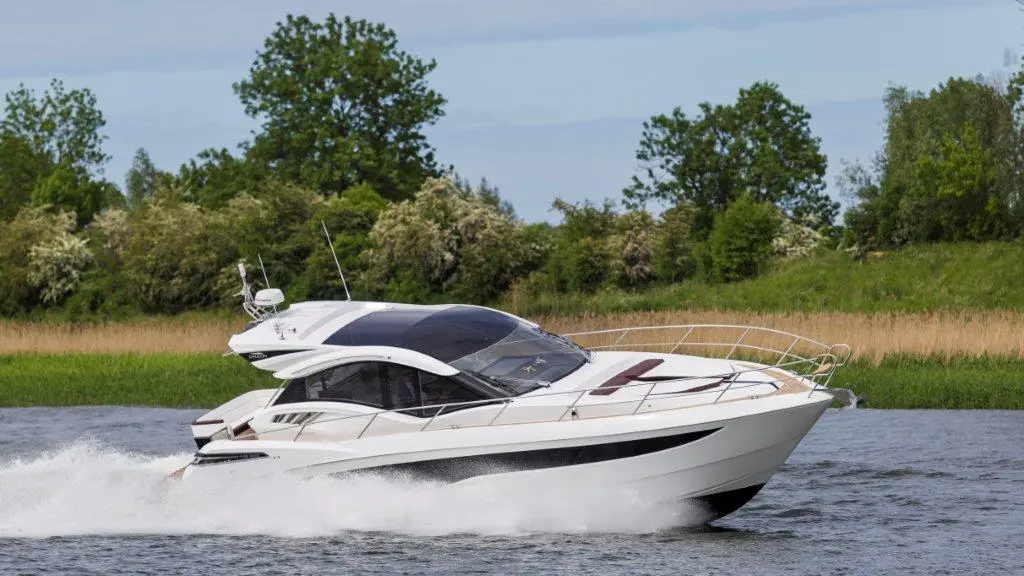 A sleek white motor yacht speeds across a calm river, surrounded by lush green trees and reeds under a clear blue sky.