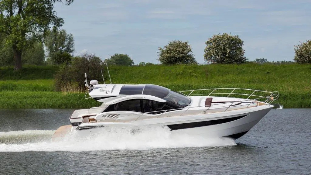A sleek motor yacht cruises on a calm river, creating a wake, with lush greenery in the background under a blue sky.