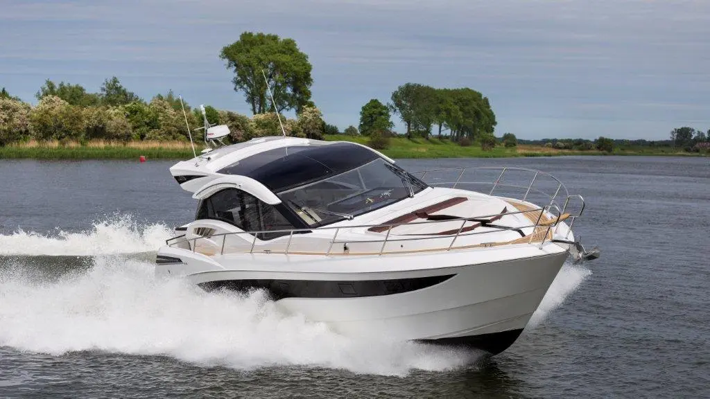 A sleek white motorboat navigates a calm waterway, creating splashes as it moves past green trees and grasslands.
