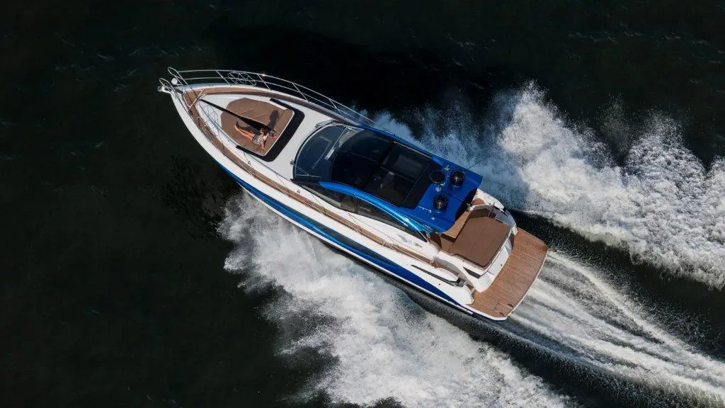 A luxury motorboat speeds across dark water, creating waves, with a sunbather lounging on the deck under clear skies.