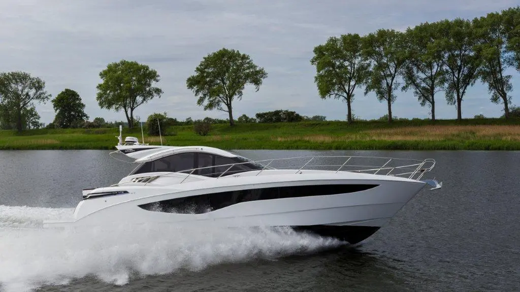 A sleek white yacht glides across a calm waterway, with lush green trees lining the shore.