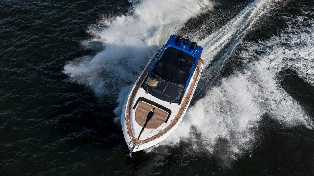 A high-speed motorboat with a blue top creates a large wake while cutting through dark water on a sunny day.