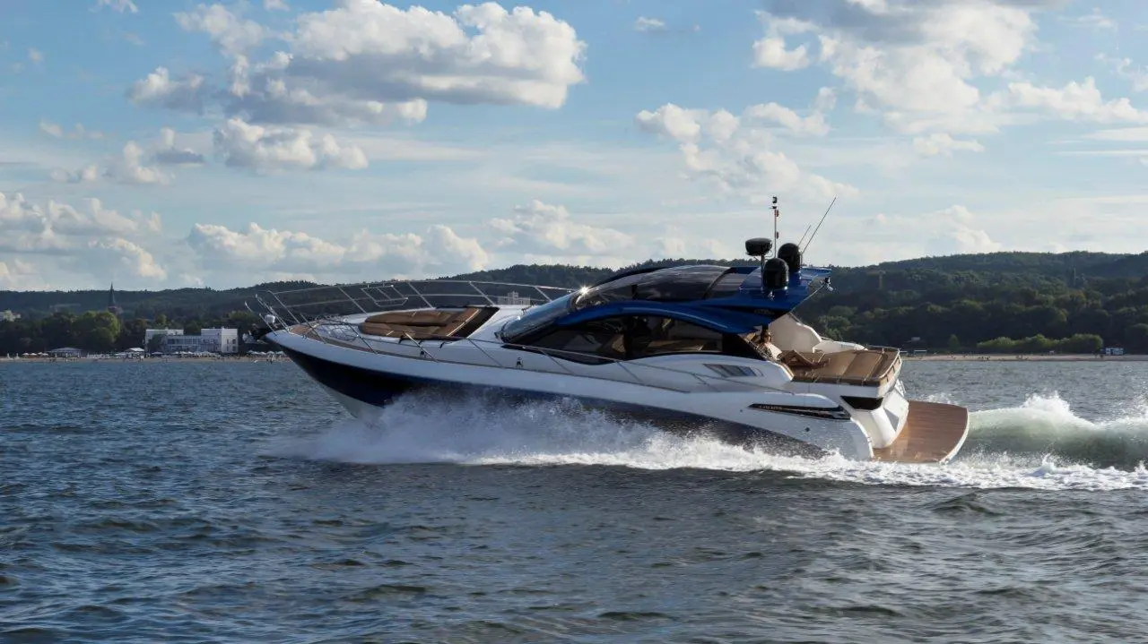 A sleek motorboat speeds across the water, leaving a wake behind, with a scenic shoreline and cloudy sky in the background.