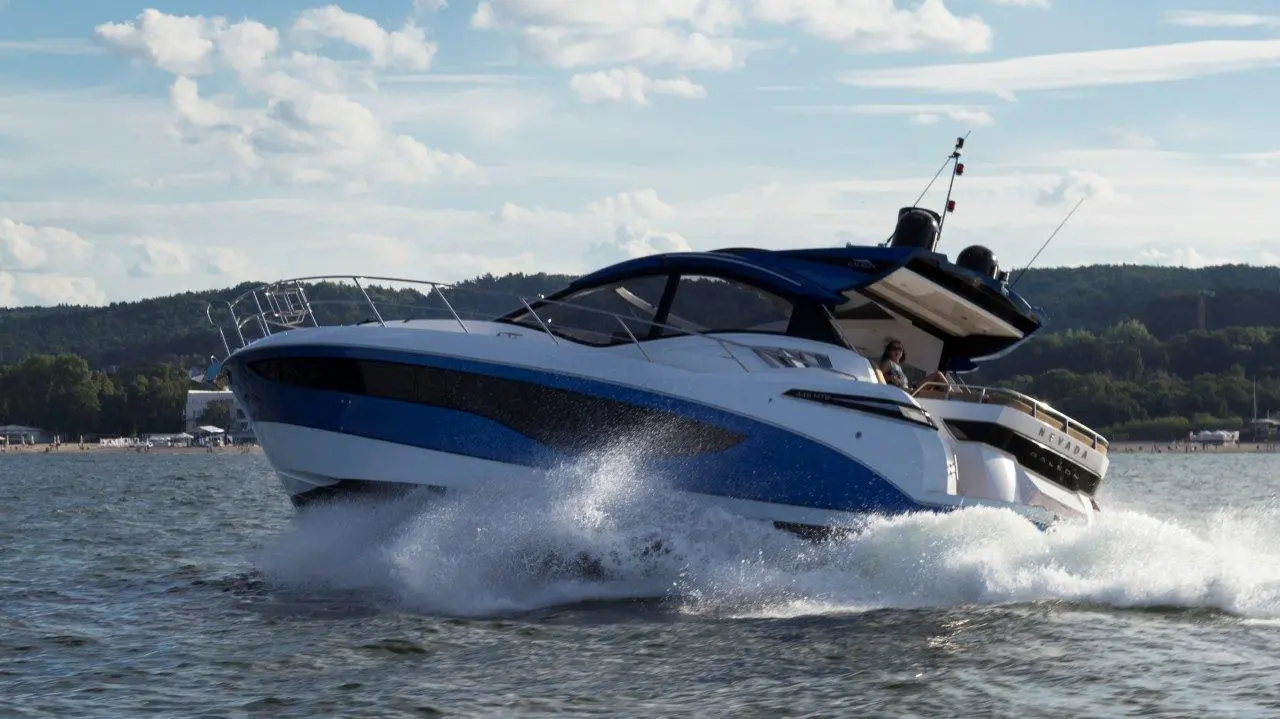 A sleek blue and white motorboat splashes through water on a sunny day, with a person at the helm and hills in the background.