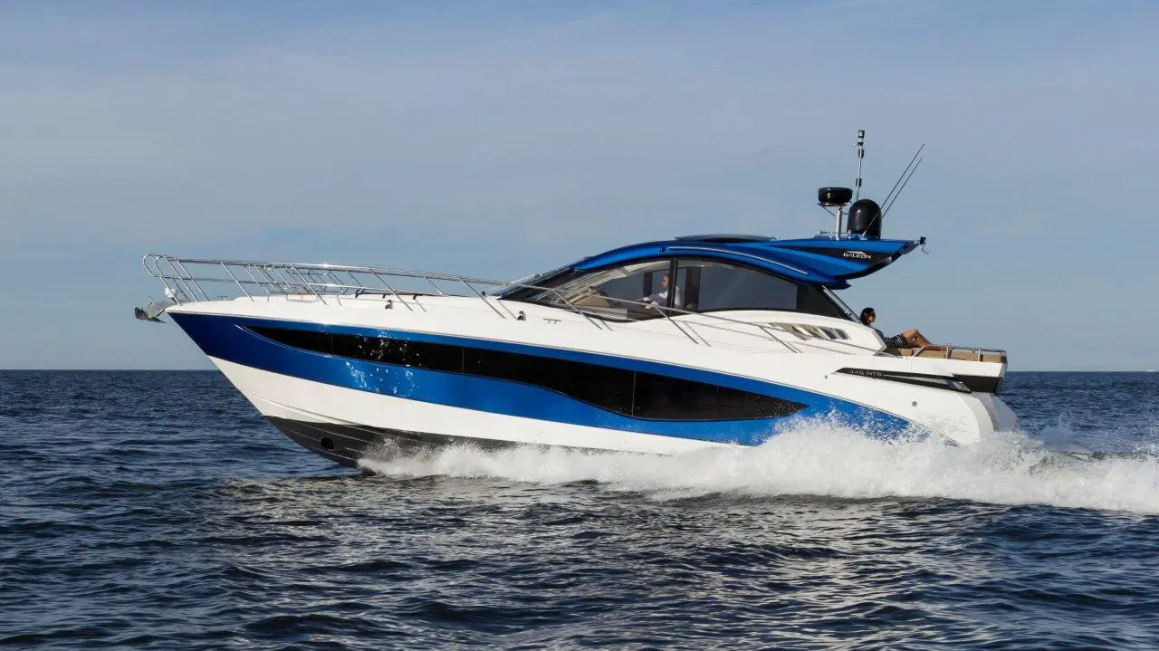 A sleek, blue and white luxury yacht speeds across the water, creating waves under a clear sky.