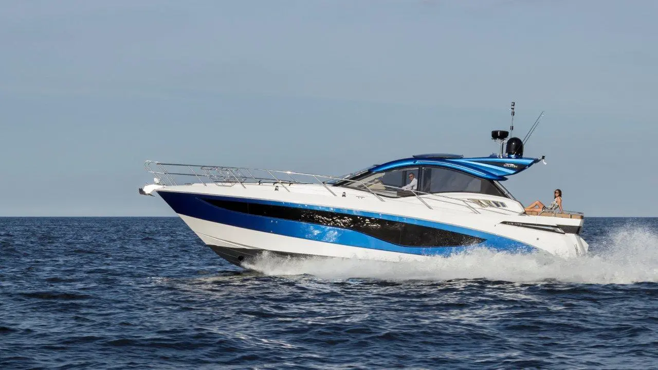 A stylish white and blue motor yacht navigates through calm blue waters, with two people on board enjoying the ride.
