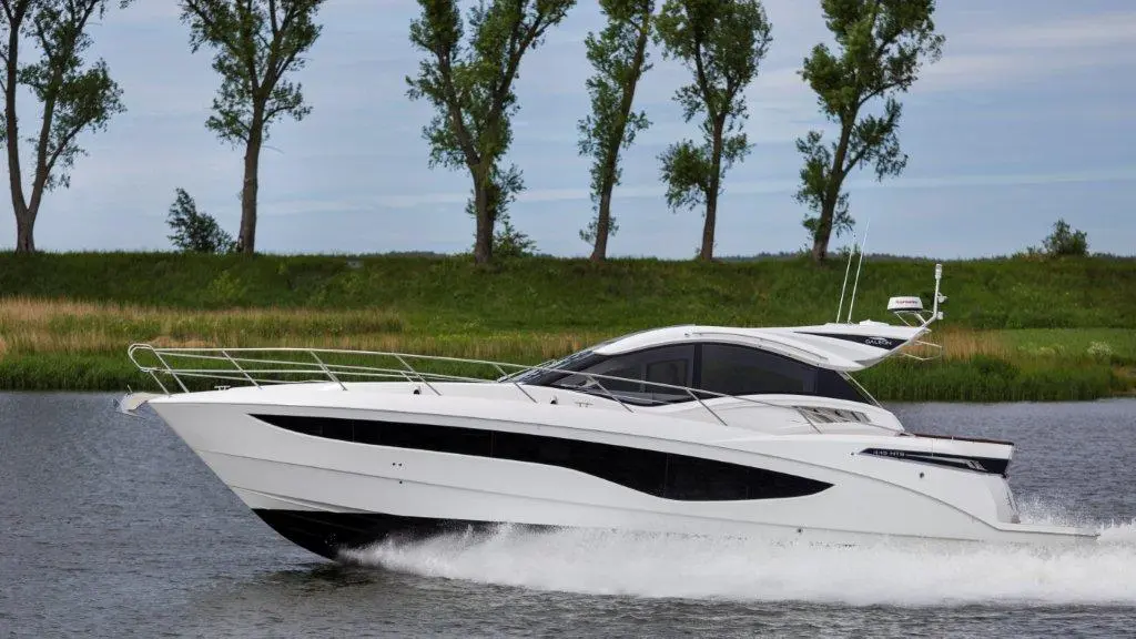 A sleek white boat speeds through calm water, with green trees lining the shore under a clear sky.