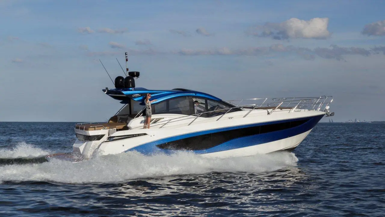 Luxury motorboat cruising on water with a person standing on deck, waving against a clear sky.