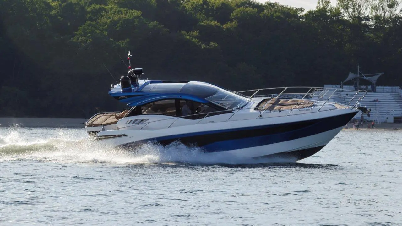 A sleek, blue and white motorboat speeds across the water, creating splashes, set against a forested shoreline.