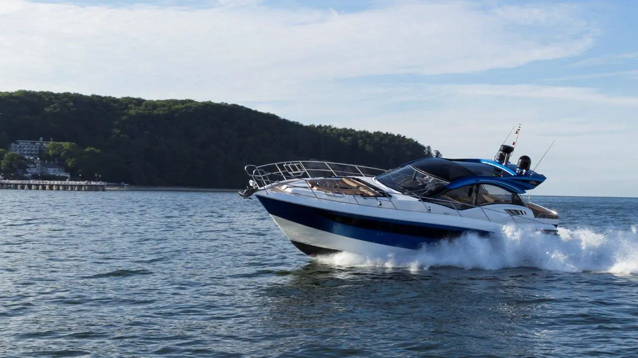 A speedboat glides across the water near a forested shoreline, creating splashes as it moves.