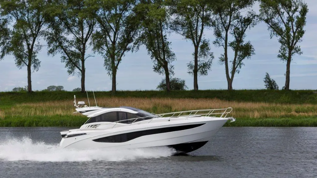 A sleek white motorboat speeds across a calm river, surrounded by green trees and grassy banks under a blue sky.